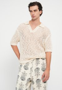 Sueter de punto de manga corta en un tono beige claro, con un escote en V y un patrón de tejido abierto; combinado con pantalones color crema con estampado floral.