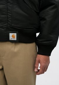 Melnā bomber jakas ar gludu tekstūru, ribotām aprocēm un apakšmalu. Uz apakšējās kreisās puses ir balts un oranžs Carhartt logo uzlīme.