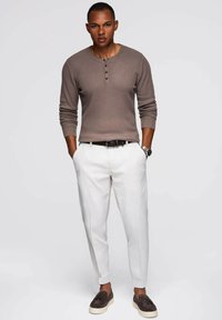 Camisa henley de manga larga marrón acanalada, pantalones blancos ajustados, mocasines oscuros, reloj de plata, manos en los bolsillos, fondo neutro.