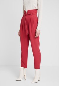 Červené kalhoty s vysokým pasem vyrobené z hladké látky s odpovídajícím páskem. Mají přední sklady a zúžené nohavice, spárované s bílými kotníkovými botami.