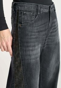 Close-up van zwarte vervaagde jeans met een donkere geblokte zijsstrijp, gedragen door een persoon met een zwart langemouwen shirt, hand rustend nabij de zak.
