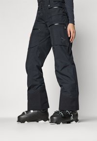 Svarta skidbyxor i vattentåligt material med sidofickor med dragkedja, förstärkta muddar och en broderad logga som accent. Har en vid bendesign.