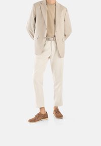 Béžový blazer, světle hnědé tričko, krémové lněné kalhoty a hnědé semišové boty. Mezi pozoruhodné prvky patří tkaný opasek a strukturovaná látka.