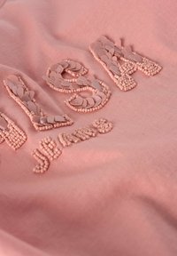 Sudadera rosa con letras texturizadas y en relieve en un tono contrastante. Tejido suave con un acabado liso. Apariencia ligeramente arrugada.