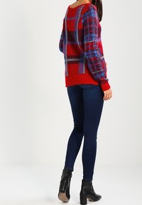 Frau trägt einen rot-blauen karierten Pullover, dunkelblaue Skinny Jeans und schwarze Stiefeletten mit Absatz, steht mit teilweise zur Seite gedrehter Rücken.