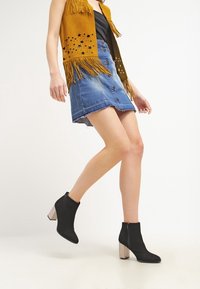 Černé kotníkové boty s lesklým blokovým podpatkem a jemným detaily zipu, v kombinaci s džínovou sukní a žlutou třásňovou semišovou vestou.