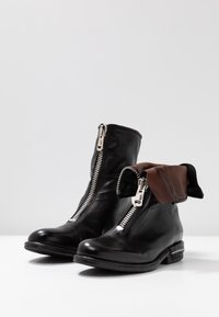 Botas de cuero negro hasta el tobillo con cremalleras plateadas, que cuentan con un puño de cuero marrón plegable y un tacón plano. Textura suave, diseño moderno.
