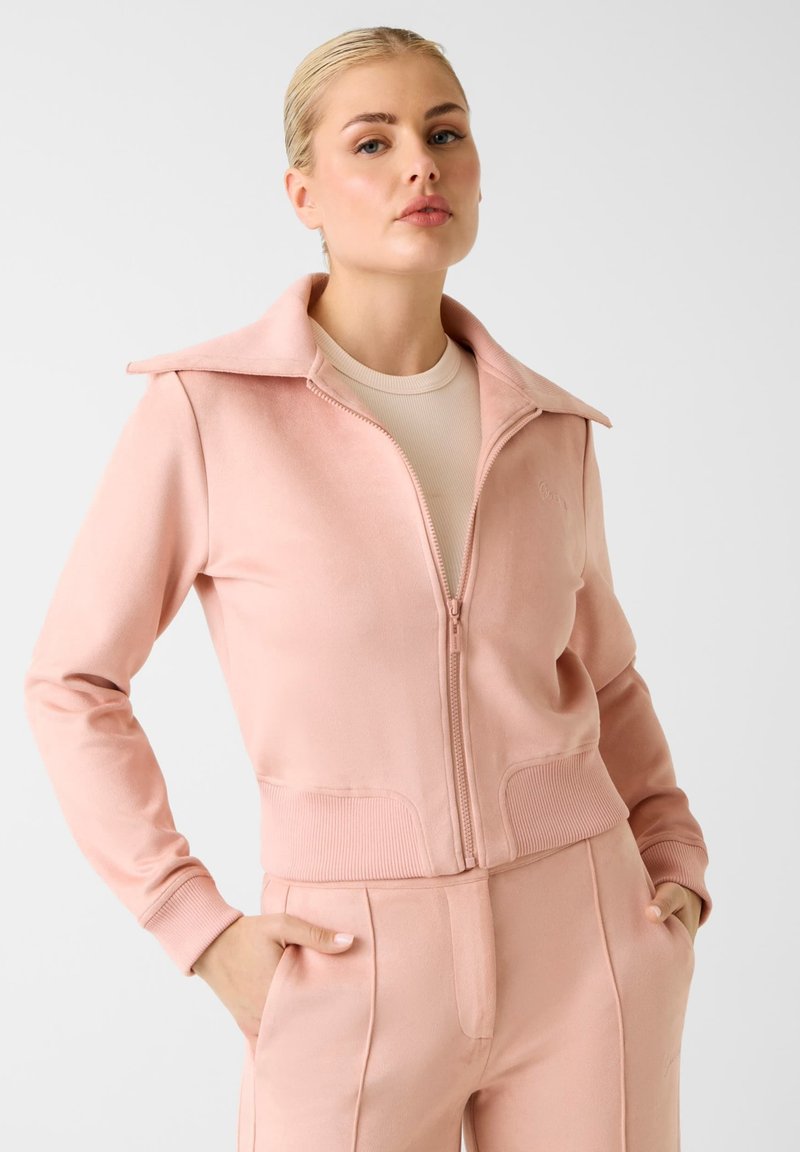 Veste rose à zip avec un large col, ourlet et poignets côtelés, fabriquée en tissu doux avec des poches latérales, associée à un pantalon assorti.