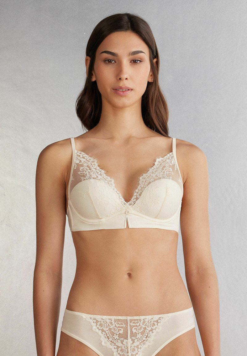 Beige beha met kantaccenten, een diepe V-hals en lichte padding. Voorzien van verstelbare bandjes en een decoratieve knoop in het midden.