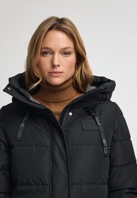 Svart puffjacka med huva, quiltad textur och märkeskännetecken. Bärs över en brun polotröja som visar en modern passform.