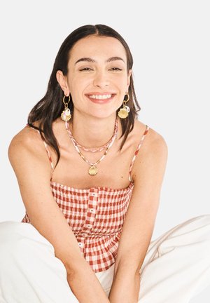 Punainen ja valkoinen ruudullinen crop-top, jossa on napit, kerroksittain simpukkakuvioinen kaulaketju ja korvakorut. Silkkiset hiukset, hymyilevä ilme.
