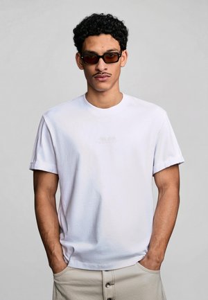 Mies pukeutunut valkoiseen t-paitaan, jonka hihat on kääritty ylös, ja beigen värisiin housuihin, kädet taskuissa. Hänellä on tummat suorakaiteen muotoiset aurinkolasit ja viikset.