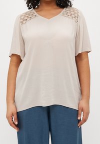 Donna che indossa una blusa beige larga con dettagli in pizzo sulle spalle e pantaloni azzurri chiari, in piedi contro uno sfondo semplice.