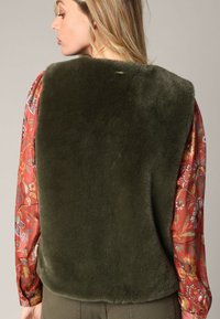 Olivgrüner Kunstfell-Westen mit weicher Textur, rundem Ausschnitt und Rückenverschluss. Kombiniert mit einem gemusterten roten Blumenhemd mit langen Ärmeln.