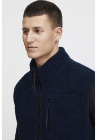 Mornarska flis prsluk s visokim ovratnikom, zatvaračem sprijeda i dva bočna džepa. Teksturirani materijal s crnim akcentima i detaljima na zatvaraču.