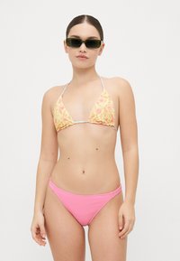 Trokutasti bikini gornji dio u žutom cvjetnom uzorku s donjim dijelom u ružičastoj rebrastoj tkanini; gornji dio ima halterske naramenice i nabran rub; model nosi tamne sunčane naočale.