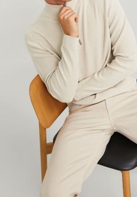 Krämfärgad ribbad tröja med hög krage, kombinerad med ljusbeige byxor. Modellen sitter på en trästol med texturerad yta.