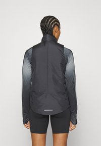 Chaqueta negra sin mangas con un cuello alto, que presenta una textura suave y un dobladillo curvado. La parte delantera es ligeramente más larga con un acabado mate.