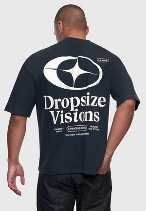 Bărbat purtând un tricou negru cu un logo alb mare și textul „Dropsize Visions”, fraze mai mici despre idei și viitor pe spate.
