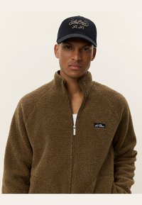 Brown fleece zip-up jacket with a textured surface and white logo patch, paired with a black cap featuring embroidered lettering.