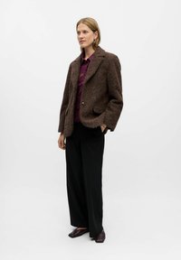 Brun tekstureret jakke med stropkraver, to knapper og sidelommer, parret med en lilla top, sorte pencilbukser og brune sko.