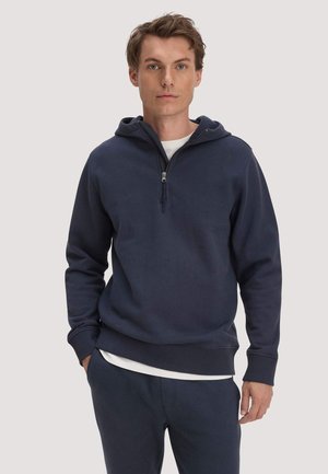 Junger Mann trägt einen marineblauen Halb-Reißverschluss-Hoodie und passende Hose mit weißem T-Shirt darunter, steht vor einem schlichten hellen Hintergrund.