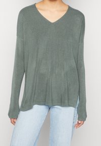 Grøn V-hals trøje lavet af blødt, tekstureret stof. Lange ærmer med en løs pasform, parret med lyseblå denim jeans.