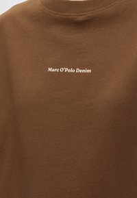 Καφέ μπλούζα με στρογγυλή λαιμόκοψη και λευκό κείμενο 'Marc O’Polo Denim' τοποθετημένο στο κέντρο του στήθους.