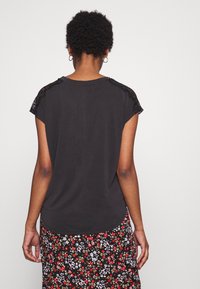 Svart t-shirt med korta ärmar, prydd med spetsdetaljer på axlarna. Mjuk tyg med rundad nederkant, buren över en blommönstrad kjol.