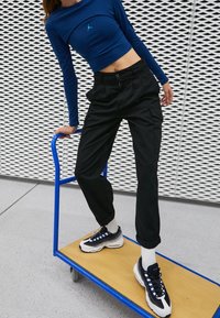 Modrý dlouhý rukáv zkrácený top s J značkou, černé cargo kalhoty, bílé ponožky a černé tenisky s modrými akcenty, na dřevěném vozíku.