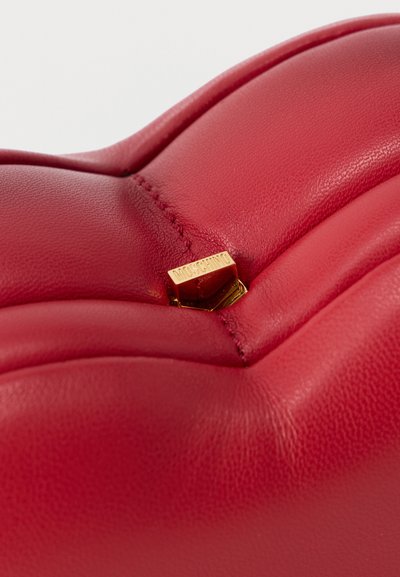 Gros plan sur le détail d'un sac à main en cuir rouge avec une plaque logo Moschino dorée au centre de la couture.