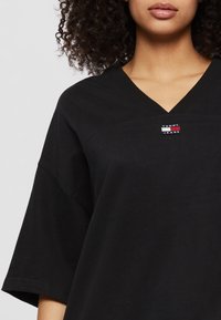 T-shirt nera a V con maniche corte e vestibilità comoda. Presenta un piccolo logo "Tommy Jeans" rosso, bianco e blu sul petto. Morbido tessuto in cotone.