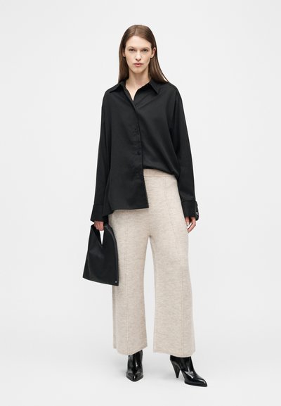 Γυναίκα με φαρδιά μαύρη πουκάμισα με κουμπιά, μπεζ πλεκτό παντελόνι με φαρδύ πόδι, μαύρα μυτερά μπότες, κρατώντας μια μαύρη τσάντα χειρός σε απλό φόντο.