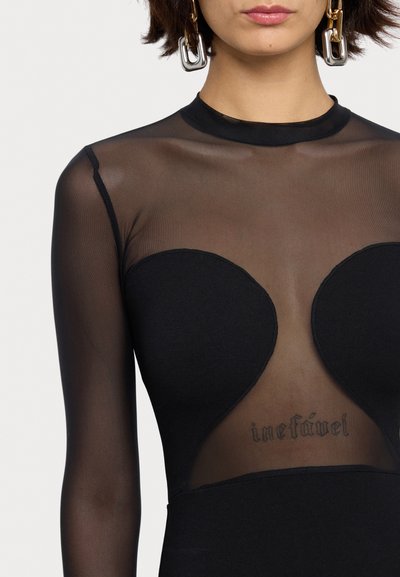 Černé priléhavé šaty s dlouhými průhlednými krajkovými rukávy a vykrojeným živůtkem. Viditelný výrazný tetování "inefável" pod krajkou. Sametově hladká textilie.