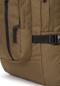 Sac à dos en toile de couleur kaki foncé, avec une poche latérale zippée, une sangle en sangle noire et une boucle en plastique, doté d'un tissu texturé et durable.