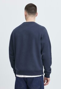Marineblauer Pullover mit langen Ärmeln, Rundhalsausschnitt und gerippten Bündchen. Weicher, strukturierter Stoff mit einem lockeren Schnitt, getragen über einem weißen T-Shirt.