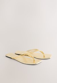 Goldene flache Sandalen mit minimalistischem Design, die dünne Riemen und ein verknotetes Detail aufweisen. Das Fußbett ist glatt und die Sohle schwarz.