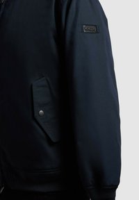 Manche et côté de veste bleu marine foncé avec poche à rabat à boutons-pression et patch rectangulaire noir portant l'étiquette "KHUJO" sur le haut du bras.