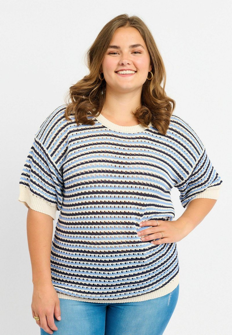 Femme souriante aux cheveux bruns ondulés portant un haut en tricot rayé bleu, blanc et noir à manches courtes et un jean bleu clair, posant avec la main sur la hanche.