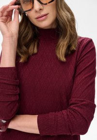 Kobieta w okularach przeciwsłonecznych w muszelkowym wzorze i długim burgundowym topie o teksturze z wysokim dekoltem, podniesioną ręką w pobliżu twarzy.