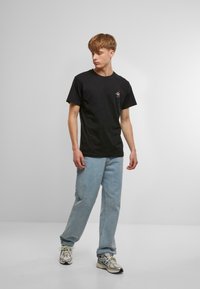 Black cotton t-shirt with short sleeves, features a small embroidered design on the chest. Worn with light blue denim jeans and silver sneakers.