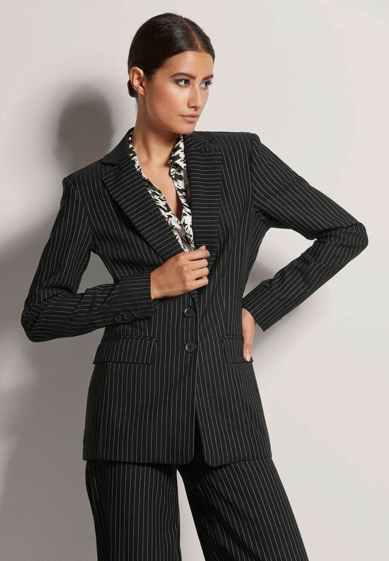 Schwarzer, gestreifter Blazer mit taillierter Passform, mit Reverskragen und Zwei-Knopf-Verschluss. Dazu wird eine gemusterte Bluse darunter getragen.