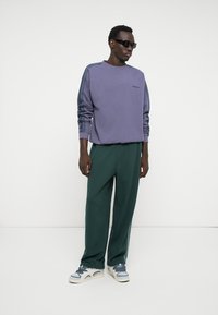 Purple sweatshirt with side striped accents, paired with loose green trousers and white sneakers. Model wearing dark sunglasses.