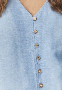 Lyseblå linned blouse med V-hals og syv træknapper nedad foran. Stoffet har en struktureret, let blank finish.
