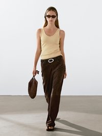 Gul ribbet tanktop, brune ruskinds brede bukser og en brun mesh håndtaske. Tilbehør inkluderer mørke solbriller og et flettet pandebånd.