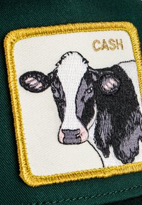 Izšūts plāksteris ar melnbaltu govs attēlu uz bēšīgas fona, ar zelta diegu roku apvilkums. Uzraksts "CASH." Plāksteris ir piestiprināts pie zaļas auduma.