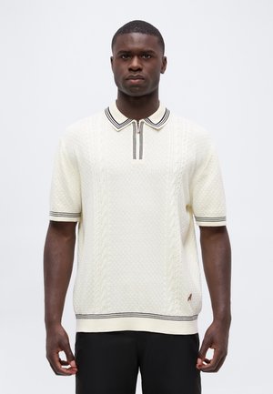 Homme portant un polo blanc texturé à manches courtes avec des rayures noires sur le col, les poignets et l'ourlet, sur un fond clair uni.