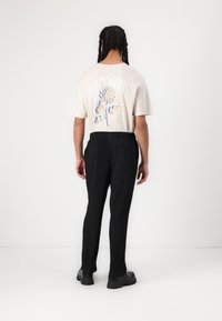 Beige bomulds t-shirt med blomster grafik og "Giftig" tekst på ryggen, parret med sorte strukturerede, plisserede bukser og kraftige sorte sko.