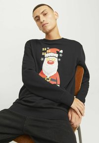 Sort sweatshirt med en farverig juleskikkelse, der bærer en rød hat, har en hvid skæg og gule stjernebriller. Ribbet manchetter og kant.