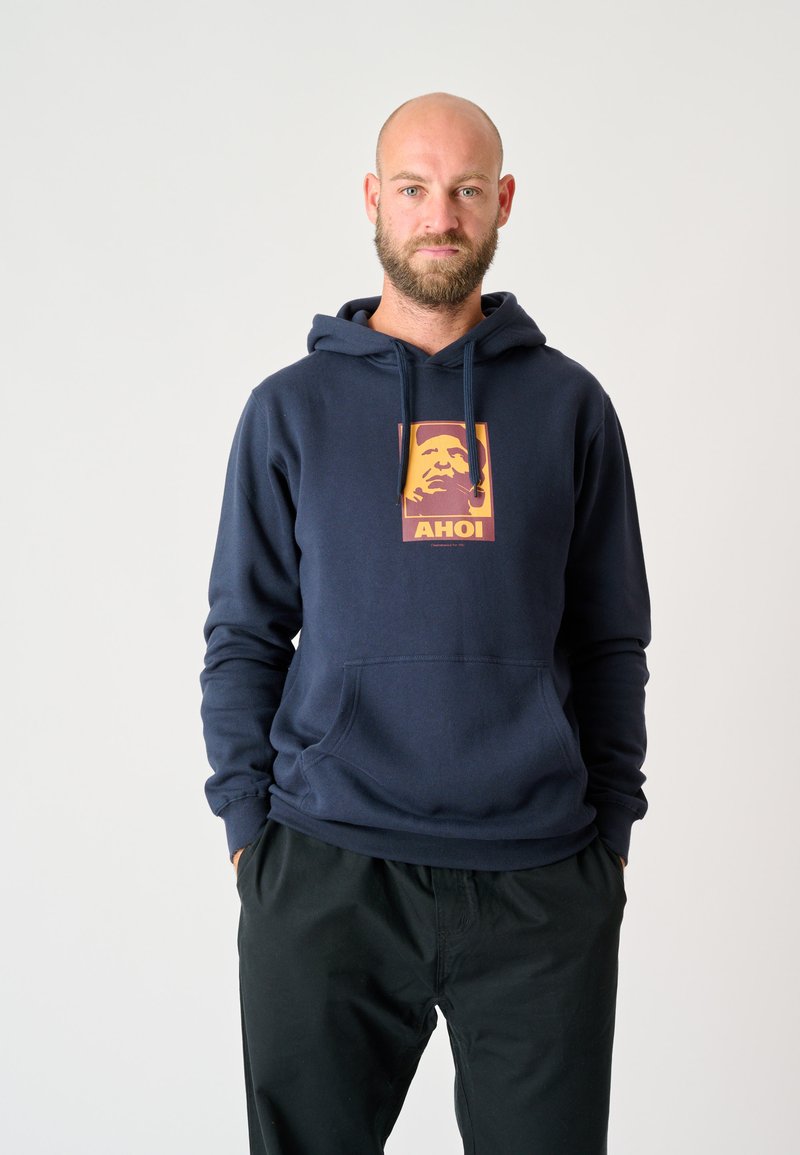 Navy-blauer Baumwoll-Pullover mit Fronttasche, ausgestattet mit einer quadratischen Grafik, die eine Illustration sowie den Text "AHOI" in orange- und gelbtönen zeigt.